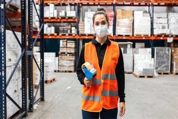 Female warehouse worker wearing protective face mask and reflective vest holding a labeling machine. Woman working in a warehouse during covid-19 pandemic.